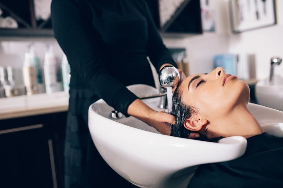 A woman relaxes at a salon while a stylist, dressed in black, gently washes her hair over a white sink. Various hair products are blurred in the background.