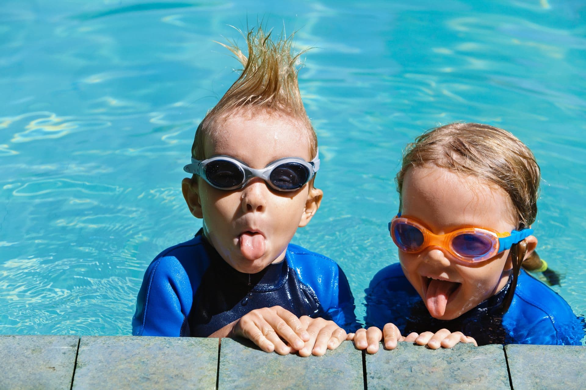 Two children in swim goggles and wetsuits stick out their tongues playfully at the edge of a bright blue pool, conveying a fun, carefree mood.