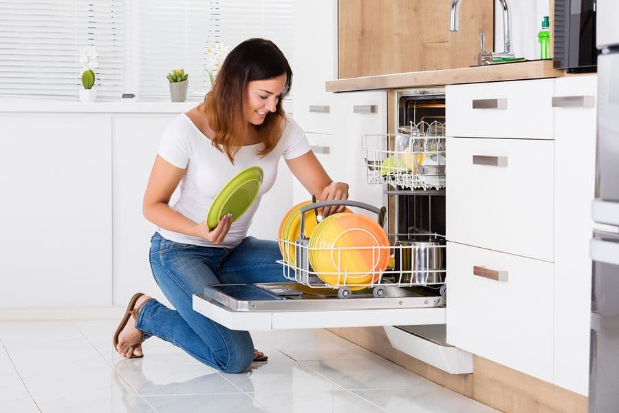 A woman kneels in a modern kitchen, unloading colorful plates from an open dishwasher. She smiles, conveying a sense of contentment and efficiency.