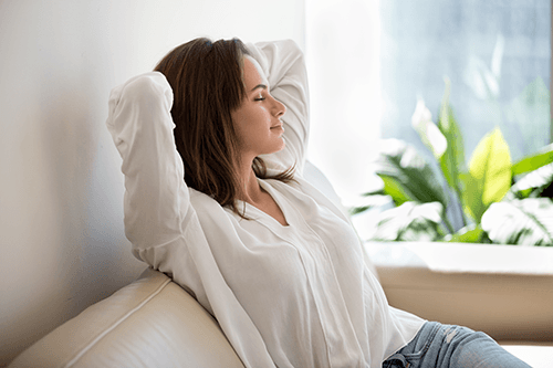 A person leans back on a couch with arms crossed overhead.