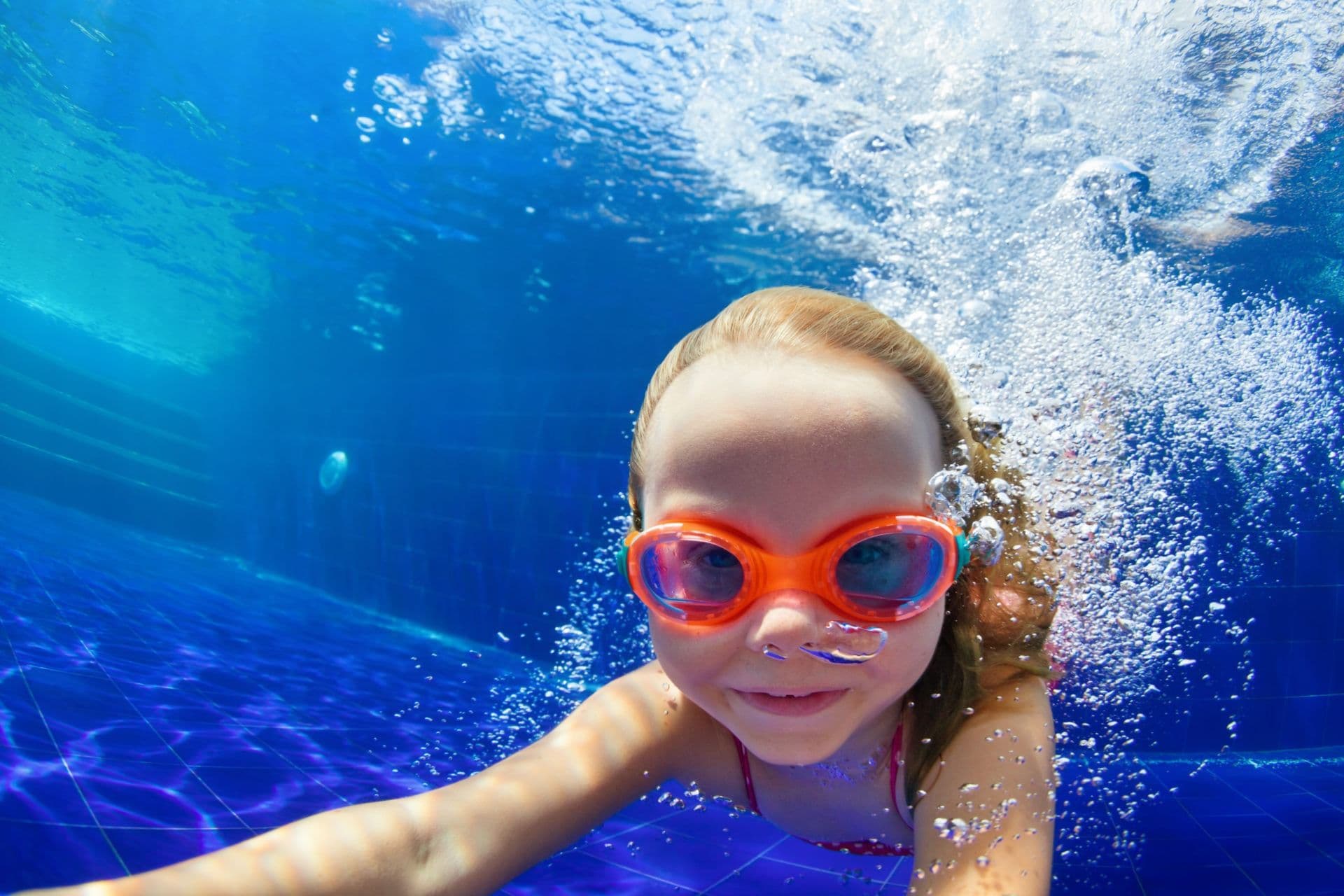 Young girl underwater in a pool with orange goggles, smiling and surrounded by bubbles. The blue water creates a joyful and playful atmosphere.