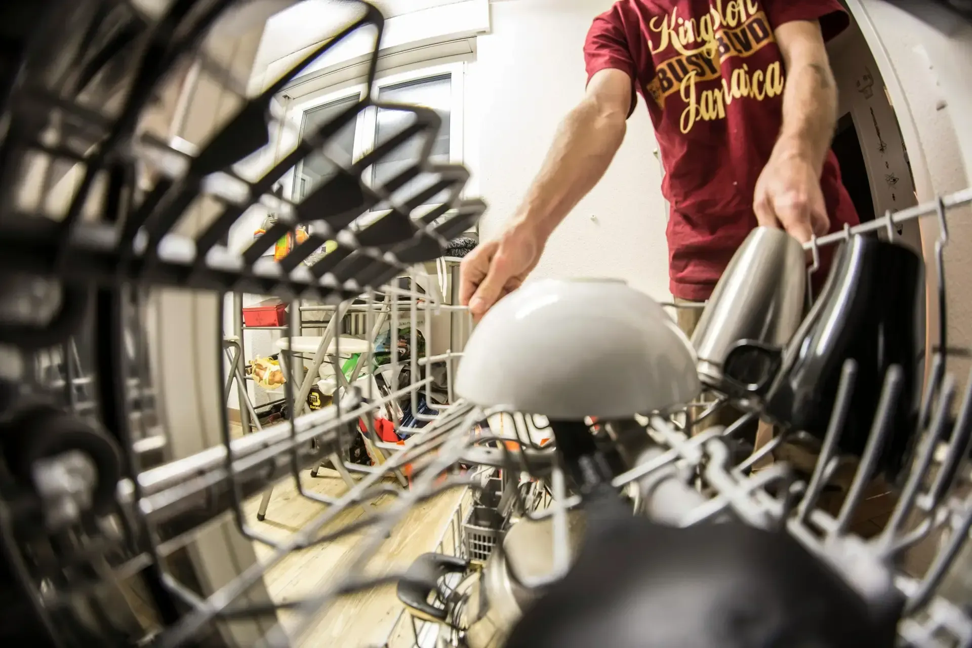 View from inside a dishwasher as a person in a red shirt loads dishes. The focus is on their hands arranging bowls and cups.