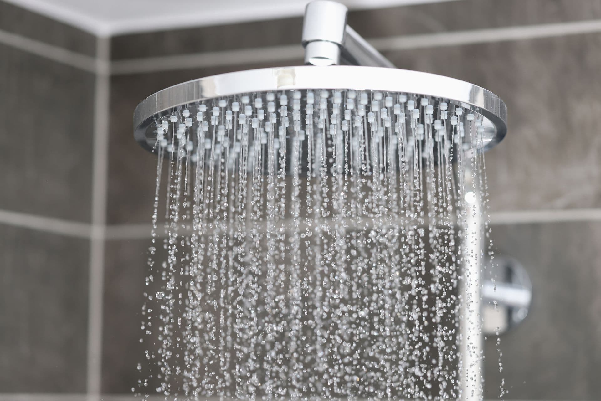 A chrome showerhead with running water against gray tiled walls, creating a refreshing and calming atmosphere. Water droplets are clearly visible.